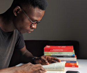 Image shows young Black man from the waist up, wearing a T-shirt and eyeglasses, while reading a paperback book. A stack of 7-8 additional books sits at his left elbow.